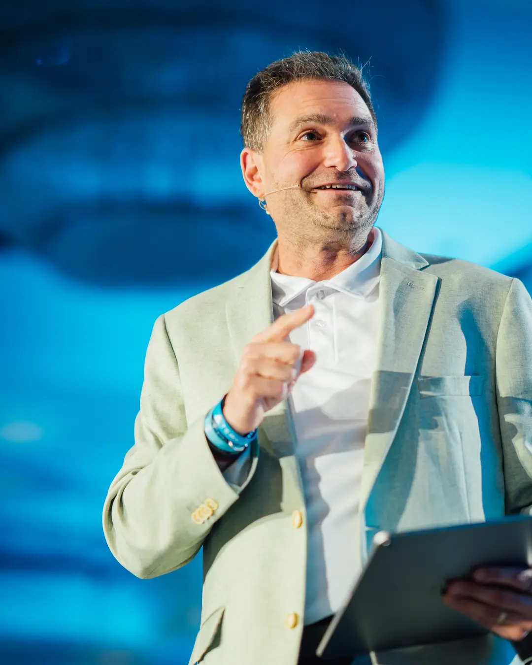 Brad Holcman speaking on stage, gesturing toward the audience under blue stage lighting.